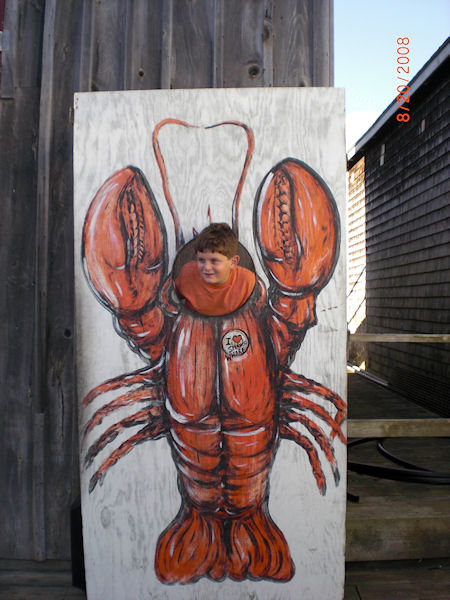 A child pokes his head out of an opening in a painted wooden board. The board depicts a giant main lobster.